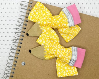 May include: Two yellow pencil-shaped hair bows with a white floral pattern and pink and silver trim. The bows are on a brown notebook with a spiral binding.