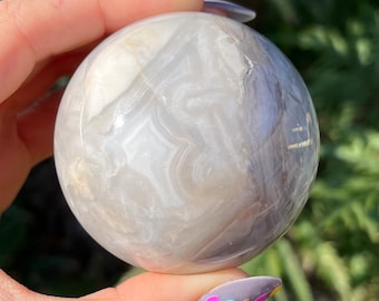 May include: A white and grey agate sphere with swirling patterns. The sphere is smooth and polished, and it is being held in a hand.