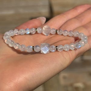 May include: A clear, beaded bracelet with a mix of round and faceted beads. The bracelet features a central, geometric-shaped bead and is strung on a flexible cord. The beads have a subtle, iridescent quality.