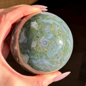 May include: A green, blue and white agate sphere with a polished finish. The sphere has a swirling pattern of colours and is a natural stone.