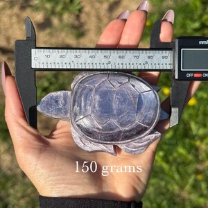 May include: A carved, lavender-coloured turtle figurine is held with a digital caliper. The figurine weighs 150 grams. The background is blurred green grass and yellow flowers.