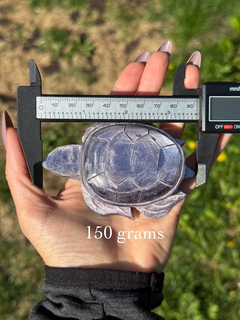 May include: A carved, lavender-coloured turtle figurine is held with a digital caliper. The figurine weighs 150 grams. The background is blurred green grass and yellow flowers.