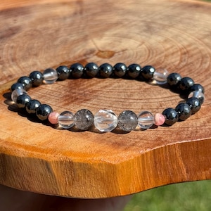 May include: A beaded bracelet featuring alternating dark gray hematite beads and clear quartz beads. The center of the bracelet includes clear quartz, pink, and gray-toned beads. The bracelet is displayed on a wooden surface.