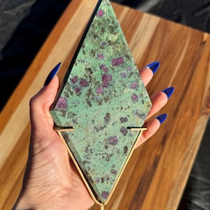 May include: A green and pink ruby zoisite crystal point on a gold metal stand. The crystal is a triangular shape with a rough, natural texture.