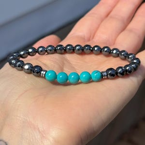 May include: A beaded bracelet featuring alternating dark gray faceted beads and turquoise blue round beads. The bracelet is held in an open hand, showcasing the contrast of colors and textures. A simple, elegant piece of jewelry.