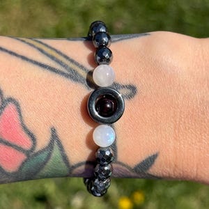 May include: A beaded bracelet featuring a central dark red stone surrounded by a silver-colored ring. The bracelet includes faceted black beads, and round white and pink beads. The bracelet is shown on a person's wrist.