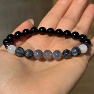 May include: A beaded bracelet featuring a combination of black, gray, and white gemstones. The bracelet is composed of polished, round beads in varying sizes. The black beads are interspersed with gray and white beads, creating a contrasting color scheme. The bracelet is displayed on a hand.