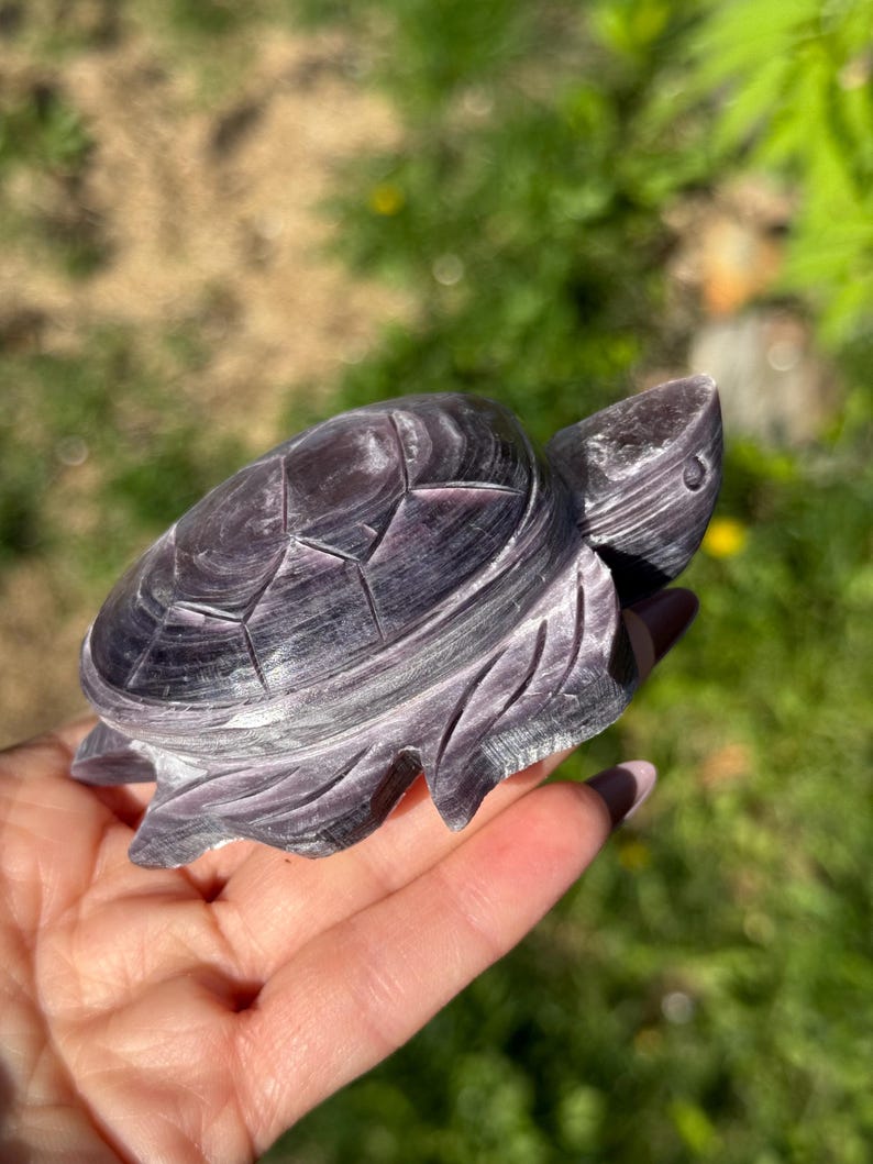 May include: A carved purple gemstone turtle figurine. The turtle has detailed shell patterns and carved flipper designs. The figurine is held in a hand against a blurred green background.