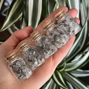 May include: Five small glass bottles with cork tops filled with clear quartz crystals. The bottles are held in a hand.