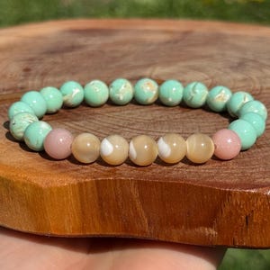May include: A beaded bracelet featuring a mix of colors. The bracelet has round beads in shades of light green, pink, and cream. The bracelet is displayed on a wooden surface, with a natural background.