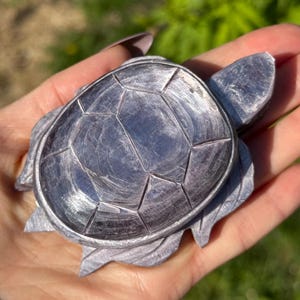 May include: A detailed, grey stone turtle figurine. The turtle has a textured shell with geometric patterns and detailed flippers and head. The figurine is held in a hand against a blurred green background.