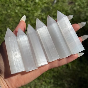 May include: Six white selenite crystal wands are held in a hand. The wands are rectangular with a pointed top. The crystals are translucent and have a striated texture. The background is blurred green grass.