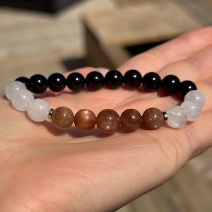 May include: A beaded bracelet featuring black, brown, and white gemstones. The bracelet has a central section of brown beads, flanked by black and white beads. The beads are round and strung together with a silver-colored metal. The bracelet is held in a hand.