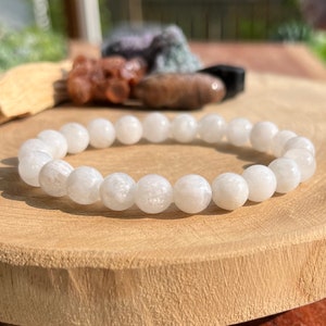 May include: White gemstone bracelet with round beads. The bracelet is made of white moonstone beads and is on a wooden surface.