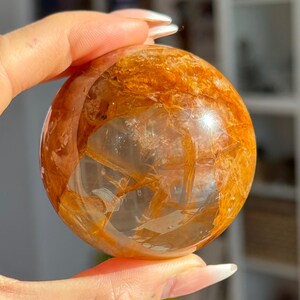 May include: A clear quartz crystal sphere with golden rutile inclusions. The sphere is held in a hand, showcasing its polished surface and the intricate patterns within the crystal. The golden strands create a striking visual contrast against the clear quartz.