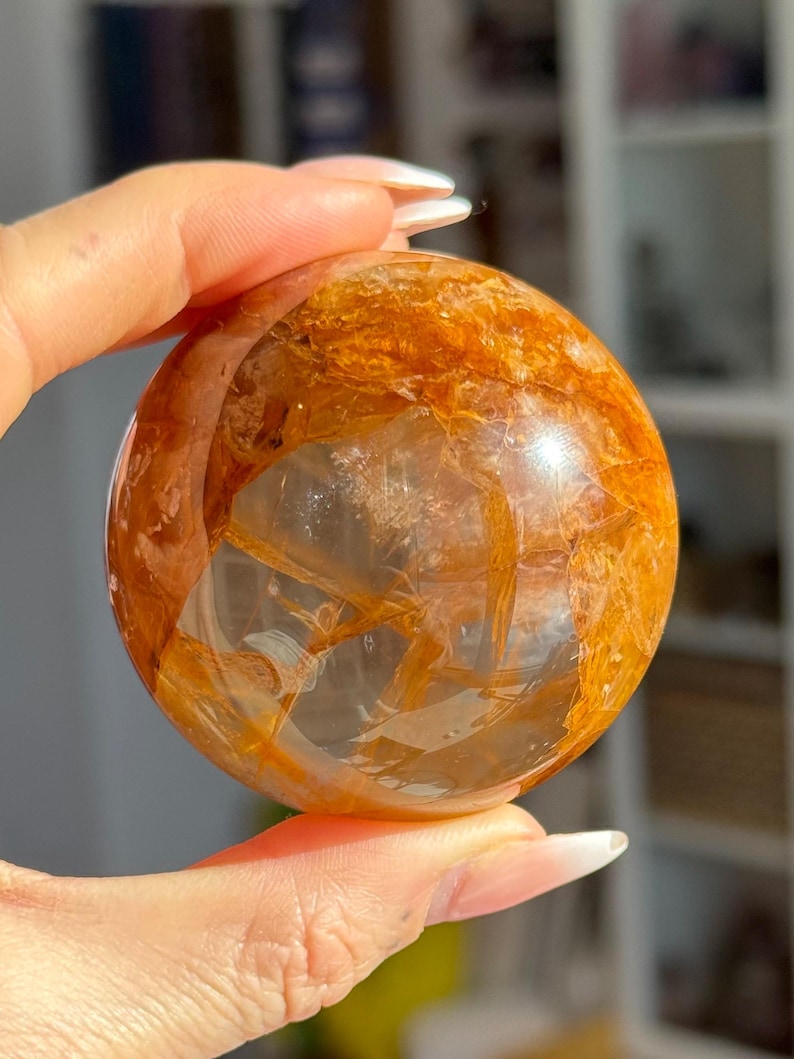 May include: A clear quartz crystal sphere with golden rutile inclusions. The sphere is held in a hand, showcasing its polished surface and the intricate patterns within the crystal. The golden strands create a striking visual contrast against the clear quartz.