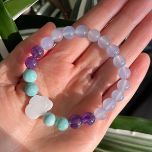 May include: A bracelet made with light blue, purple and green beads. The bracelet has a white cloud charm.