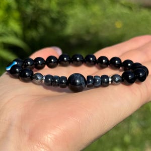 May include: Black beaded bracelet with a variety of bead shapes and sizes. The bracelet includes a large, round, dark blue bead and a small blue and white evil eye charm. The bracelet is displayed on a hand.