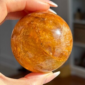 May include: A golden-orange crystal sphere with internal patterns. The polished gemstone is held in a hand, showcasing its round shape and translucent quality. The sphere's color ranges from light amber to deep orange.