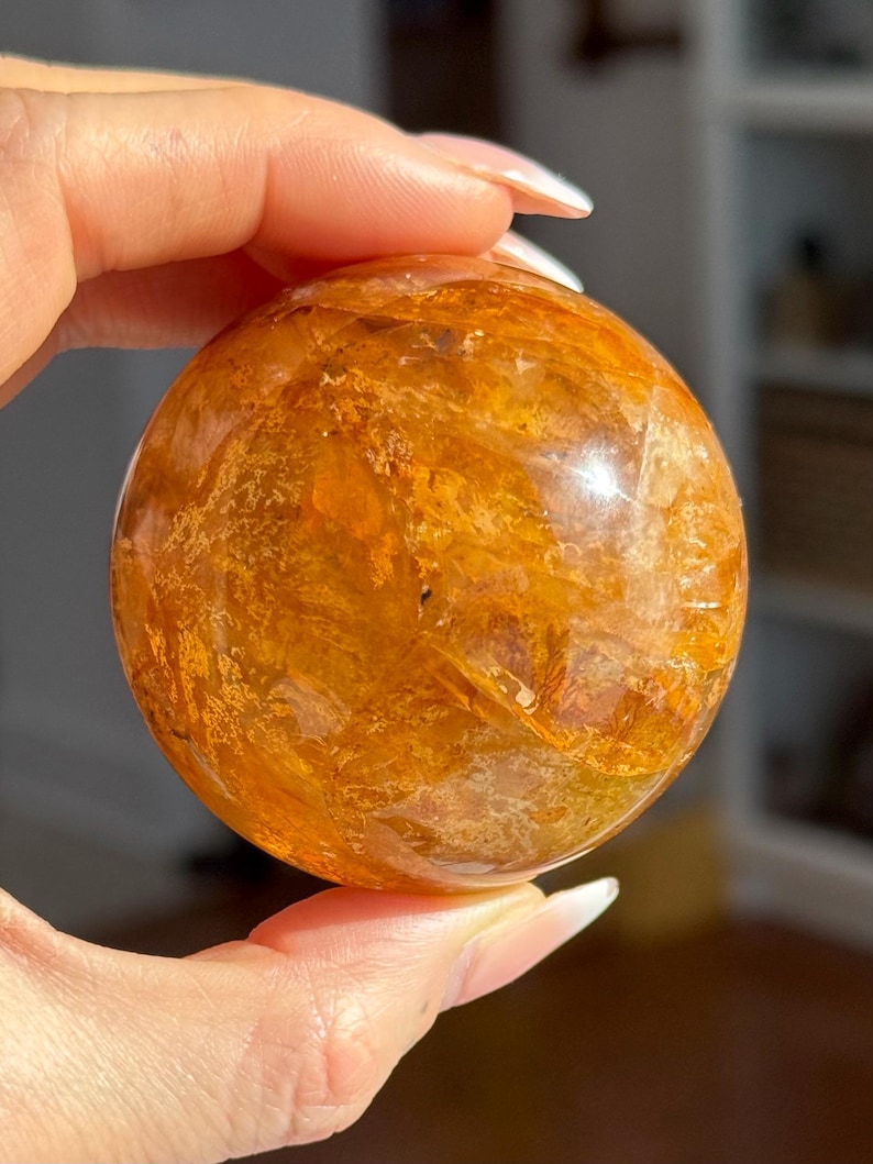 May include: A golden-orange crystal sphere with internal patterns. The polished gemstone is held in a hand, showcasing its round shape and translucent quality. The sphere's color ranges from light amber to deep orange.