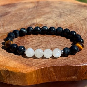 May include: A beaded bracelet featuring black, tiger's eye, and white moonstone beads. The bracelet is strung on an elastic band and is displayed on a wooden surface. The beads are approximately 0.8 cm in diameter.