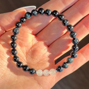 May include: A beaded bracelet featuring dark blue and black spherical beads. The bracelet includes three white, translucent beads. The bracelet is strung on a clear elastic cord.
