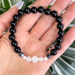 May include: A black beaded bracelet with a mix of black, white, and gray beads. The bracelet features large, glossy black beads interspersed with smaller white and gray beads. The bracelet is shown on a hand.