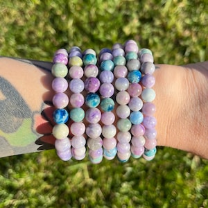 Peut inclure: Un ensemble de huit bracelets en perles de pierres précieuses de couleur lavande, bleu et vert. Les bracelets sont empilés sur le poignet d'une personne.