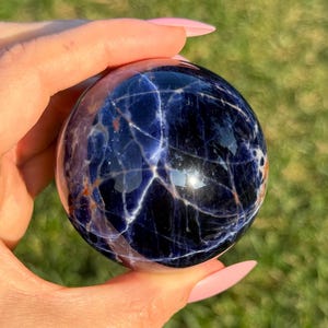 May include: A polished blue sodalite sphere with white veins and a hint of orange. The sphere is smooth and reflective.