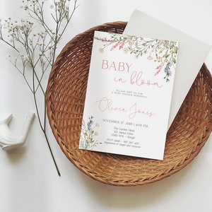 May include: A baby shower invitation with the text "Baby in Bloom" and the name "Olivia Jones" in a floral design. The invitation is on a white background, placed inside a woven basket with a white envelope.
