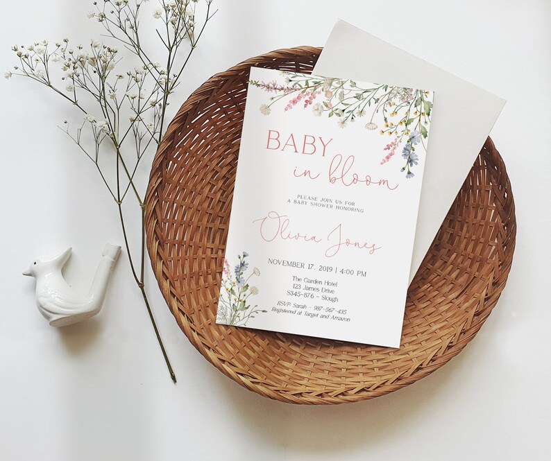May include: A baby shower invitation with the text "Baby in Bloom" and the name "Olivia Jones" in a floral design. The invitation is on a white background, placed inside a woven basket with a white envelope.