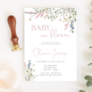 Pu&ograve; includere: Un invito per un baby shower con decorazioni floreali e la scritta "BABY in bloom". L'invito include il nome "Olivia Jones" e i dettagli dell'evento. Un timbro in legno con base dorata &egrave; a sinistra.
