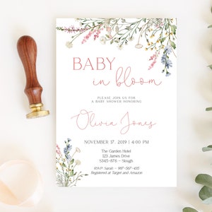 May include: A baby shower invitation with floral accents and the text "BABY in bloom." The invitation includes the name "Olivia Jones" and event details. A wooden stamp with a gold base is on the left.