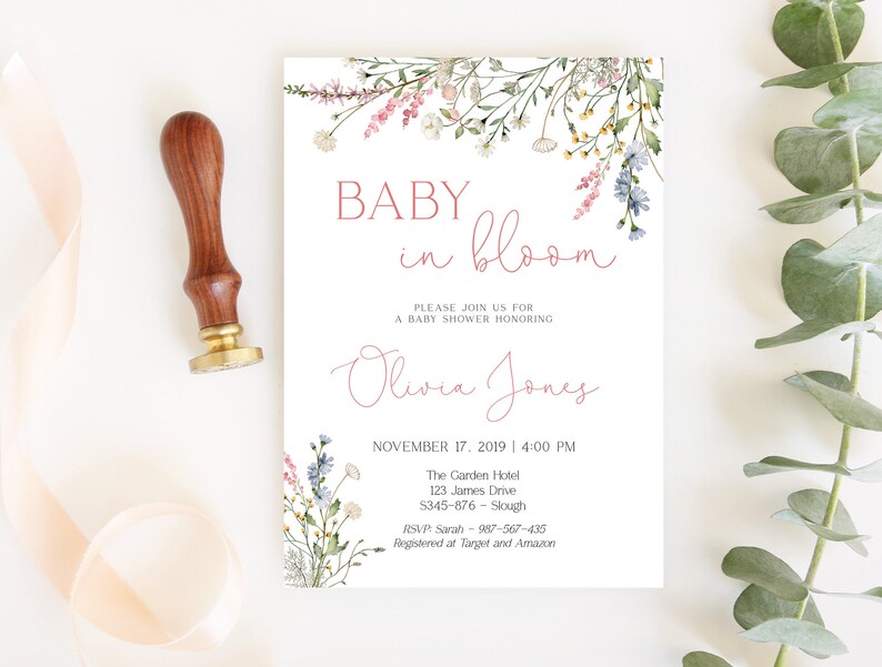 May include: A baby shower invitation with floral accents and the text "BABY in bloom." The invitation includes the name "Olivia Jones" and event details. A wooden stamp with a gold base is on the left.