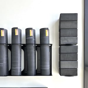 May include: A wall-mounted storage system with rolled yoga mats and stacked yoga blocks. The dark grey mats are held in a black metal rack. The blocks are also dark grey and arranged vertically, offering a space-saving solution for fitness equipment.