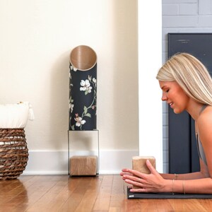 Op de afbeelding: Een vrouw beoefent yoga op een zwarte mat met een bruine yogablok. Ze draagt een grijs tanktop en heeft haar handen op de blok. De blok ligt op een houten vloer. Op de achtergrond staat een zwart-witte, bloemige cilindervaas op een zwarte metalen standaard.