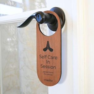May include: Wooden door hanger with the text "Self Care In Session" and "please honour the silence". The hanger is brown with black text and a black hand symbol. The text "moche" is at the bottom of the hanger.