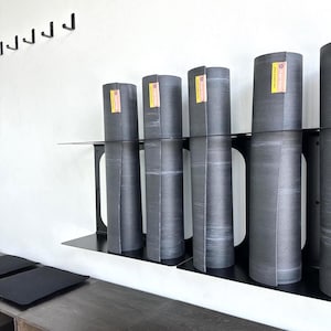 May include: Rolled-up gray yoga mats with yellow and pink labels are displayed on a black metal shelf. The mats are cylindrical and have a textured surface. The shelf is mounted on a white wall, with black hooks above.