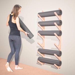 May include: A wooden yoga mat rack with five slots, each holding a rolled-up gray yoga mat. The rack is mounted on a light-colored wall. A person is holding a rolled-up gray yoga mat.