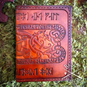 Op de afbeelding: Een rechthoekige notitieboekhoes van bruin leer met ingewikkeld reliëf vlechtwerk en een centraal drakenmotief. Het oppervlak is versierd met uitgesneden runeninscripties en donkere stiksels langs de randen, wat zorgt voor een getextureerd uiterlijk.