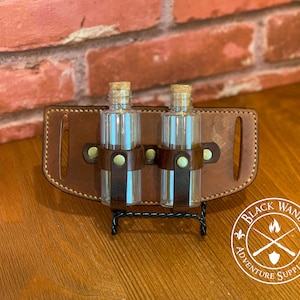 May include: A brown leather belt with two clear glass bottles with cork tops. The belt has two brass buckles and is labeled "Black Wand Adventure Supply Co."