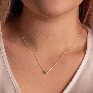 May include: A silver chain necklace with a small blue evil eye charm. The necklace is worn by a person with long blonde hair.