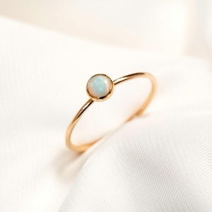 May include: A delicate gold ring featuring a small, round opal gemstone. The ring is displayed on a white fabric background.