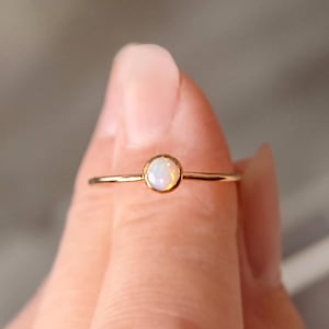 May include: A dainty gold ring featuring a small, round opal gemstone. The opal is milky white with hints of iridescent blue and green.