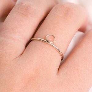 May include: A silver ring with a small circle on top. The ring is worn on a finger.