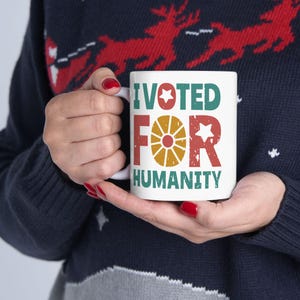 Puede incluir: Taza de cerámica blanca con el texto "I VOTED FOR HUMANITY" en verde, rojo y amarillo. La taza es sostenida por una persona que lleva un suéter azul marino con un patrón de renos rojos.