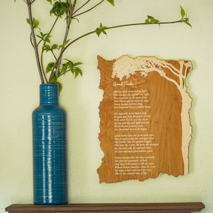 May include: A teal blue ceramic vase with horizontal etched lines holds leafy branches. To the right, a wooden plaque with the text "Good Timber" and a tree design is mounted on the wall. The plaque is made of light and dark wood.