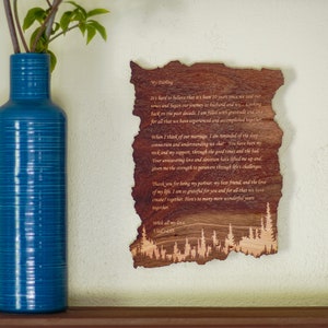 May include: A wooden plaque with a message and a forest silhouette. The plaque is made of dark wood and has an irregular shape. A blue vase with green leaves is on the left. The text is visible.