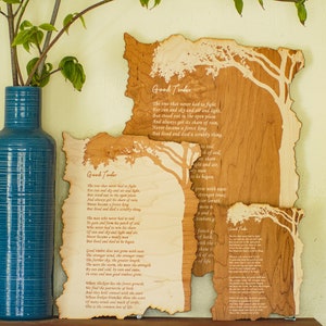 May include: Three wooden art pieces with a tree design and the poem "Good Timber" etched into the wood. A tall, blue vase with a plant is to the left of the art pieces. The wood has a natural, light brown color.
