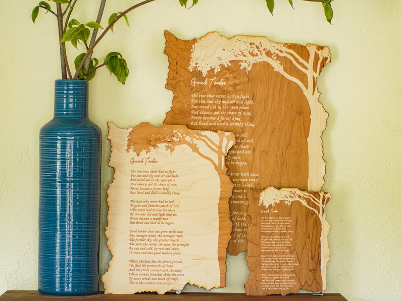 May include: Three wooden art pieces with a tree design and the poem "Good Timber" etched into the wood. A tall, blue vase with a plant is to the left of the art pieces. The wood has a natural, light brown color.